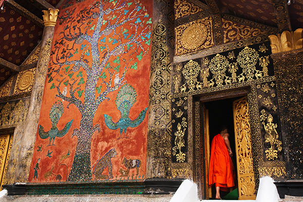 exterior of ancient temple of Wat Xieng Thong, magical stained glass on wall, young Laotian Buddhist monk stands at the door. Luang Prabang, Laos