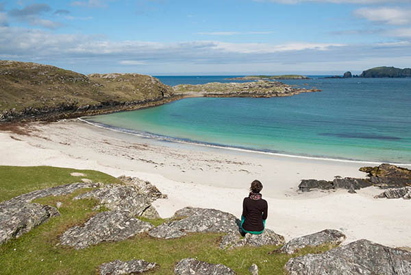 Young woman tourist sitting in front of a desert white beach with blue sea in the Isle of Lewis, Outer Hebrides, Scotland