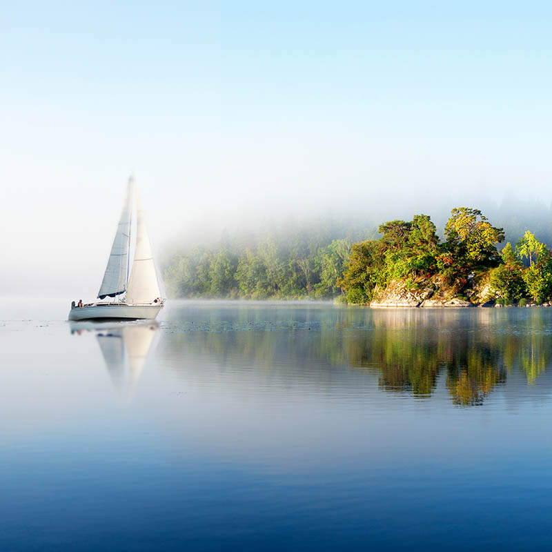 Island and yacht reflected in clear blue Scandinavian lake on foggy sunny morning