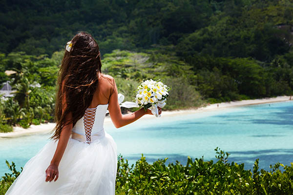 Young bride standing with a bouquet looking at a beach with blue water in the Seychelles