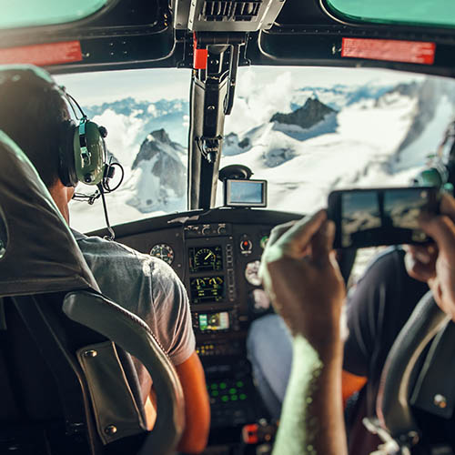 Interior of helicopter cockpit with pilot and co-pilot, and blurred tourist's arms taking photos with a mobile phone.