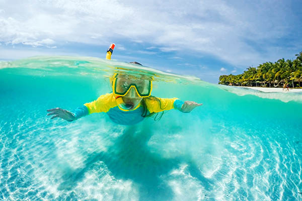 Half underwater photo of child in mask snorkeling in blue ocean water near tropical island