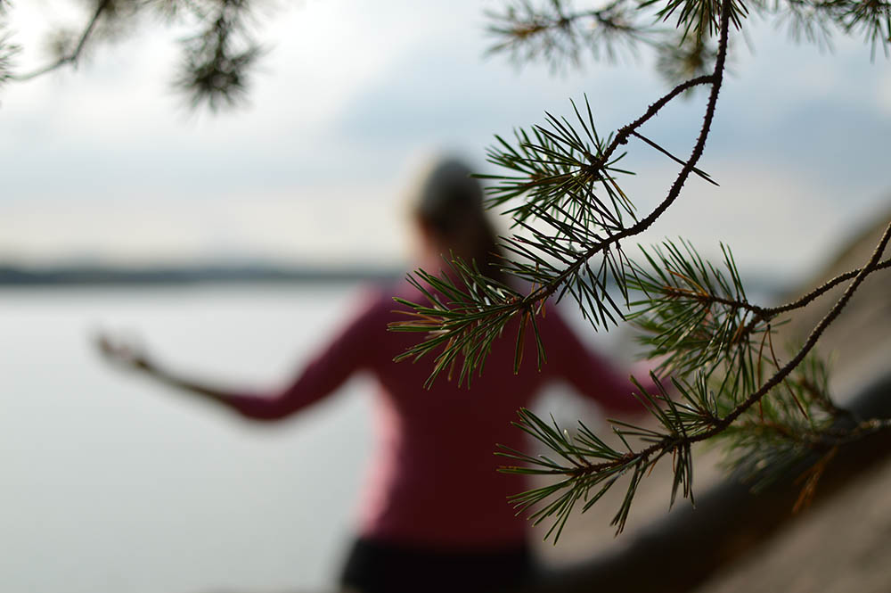 A woman in pink shirt is meditating outdoors by water. Selective focus on the tree branch in front, blurred background