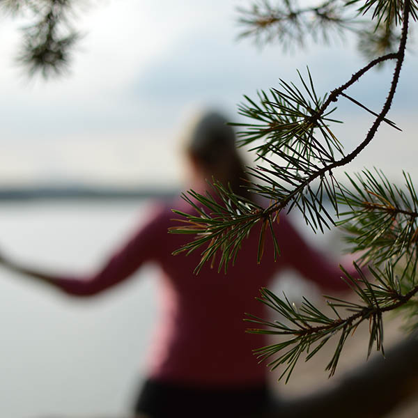 A woman in pink shirt is meditating outdoors by water. Selective focus on the tree branch in front, blurred background