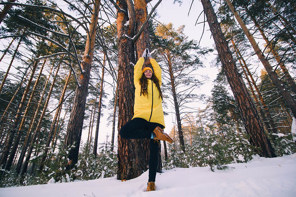 young woman in yellow jacket and hat doing yoga among snow covered trees