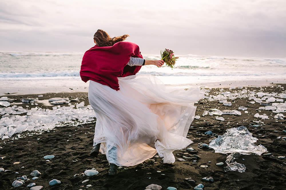 Bride in white dress with red shawl walking on a windy day on Black Sand Beach, Iceland