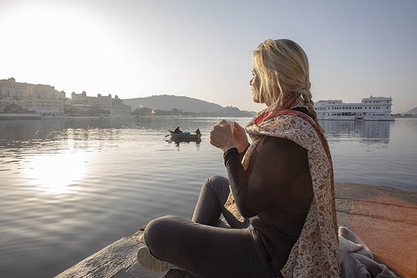 Mature woman sits on a ghat at sunrise with a hot drink with the Taj Lake Palace on Lake Pichola in the background. Udaipur, Rajasthan, India