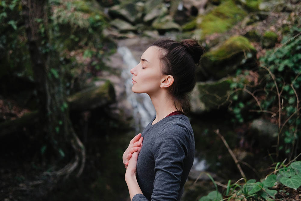 Young woman practicing breathing yoga pranayama outdoors in moss forest on background of waterfall