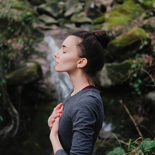 Young woman practicing breathing yoga pranayama outdoors in moss forest on background of waterfall