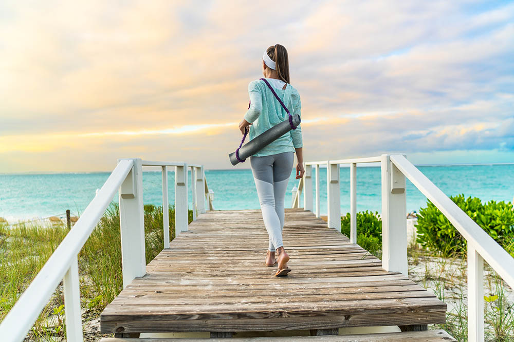 woman walking with yoga mat on beach going to outdoor meditation class at sunset