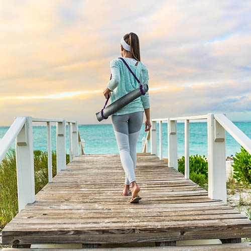 woman walking with yoga mat on beach going to outdoor meditation class at sunset