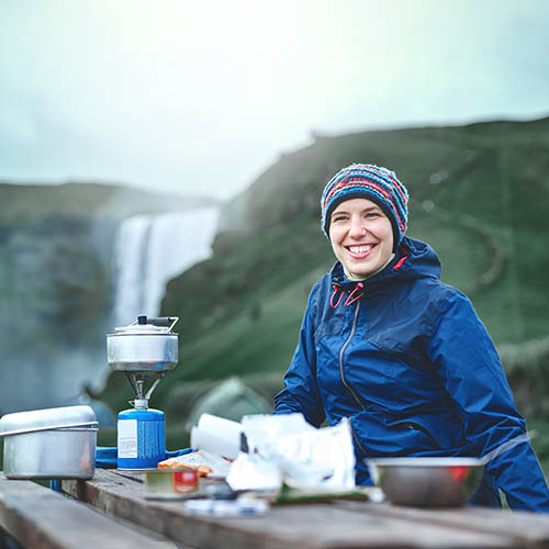 Two women preparing food on a gas stove while camping near a waterfall in Iceland