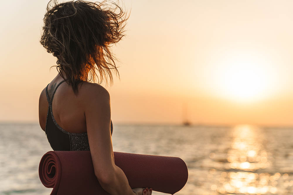 Back view of woman standing with yoga mat beside the sea in the morning