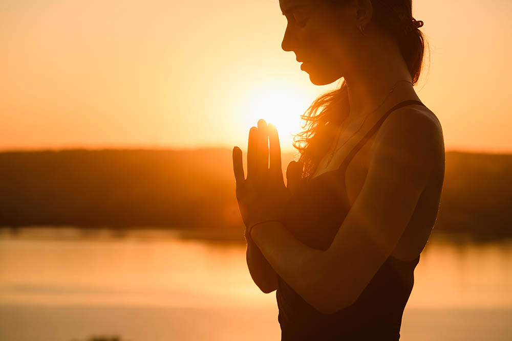 Woman in yoga meditation pose outdoors by river at sunset.